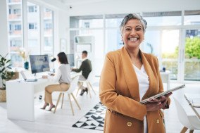 Woman in a tan jacket holding a digital pad in the office with two coworkers behind her